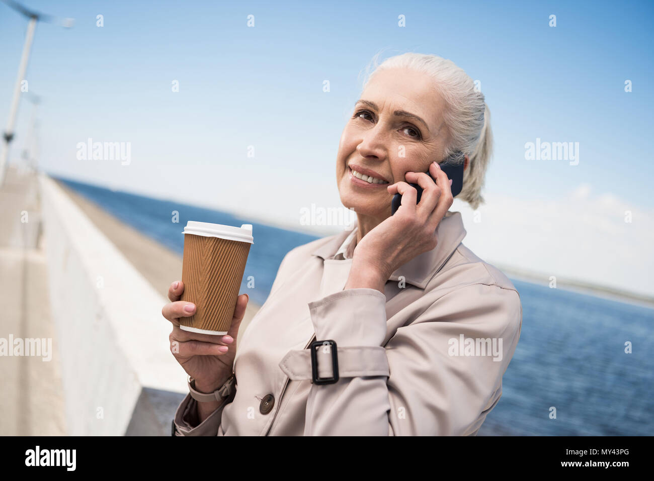 Sorridente donna anziana parlando su smartphone e azienda tazza di caffè al quay Foto Stock