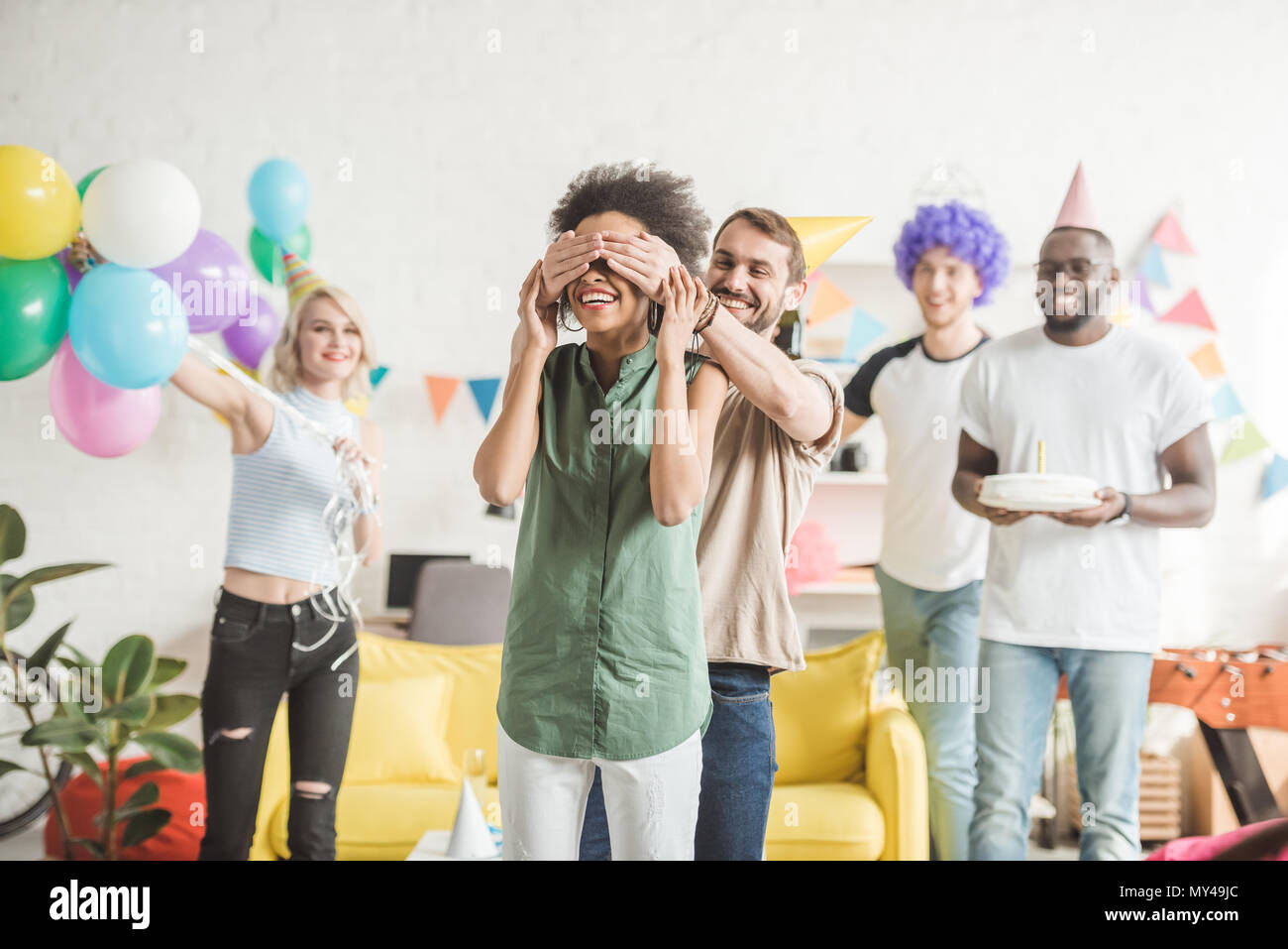 Happy amici che coprono gli occhi della giovane donna e saluto lei con una torta di compleanno in festa a sorpresa Foto Stock