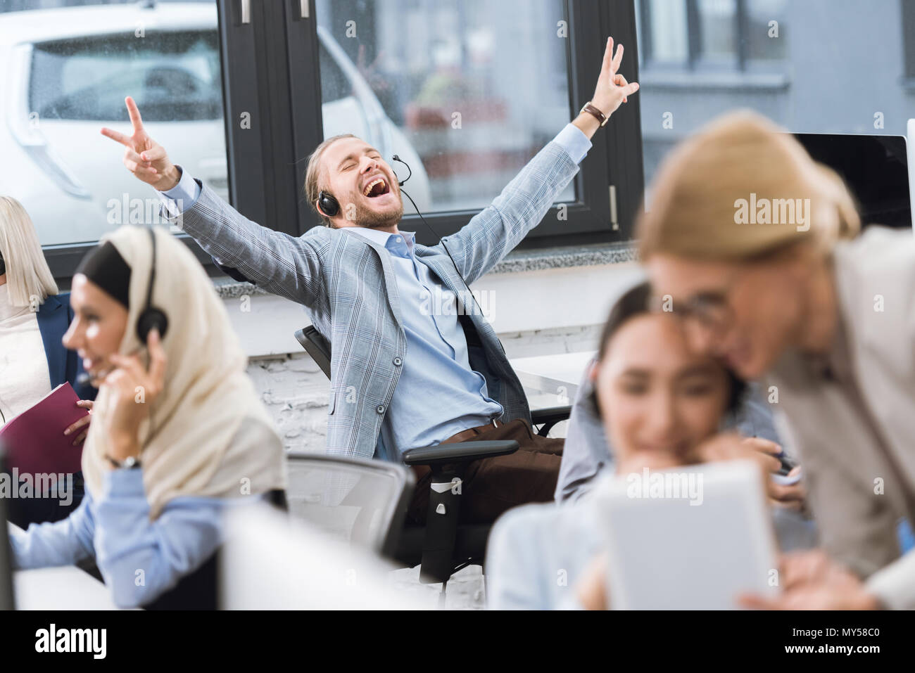 Messa a fuoco selettiva di happy businessman in cuffia che mostra segni di pace stando seduti al posto di lavoro in ufficio Foto Stock