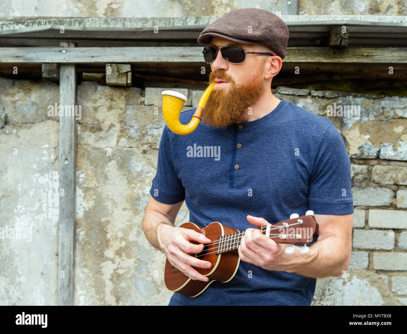 Foto di un uomo barbuto trentenne fumare una canalizzazione di grandi dimensioni e la riproduzione di un ukulele. Foto Stock