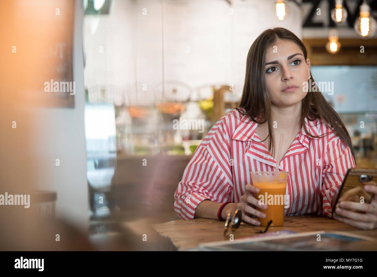 Giovane donna seduta al cafe, tenendo lo smartphone, frullato potabile Foto Stock