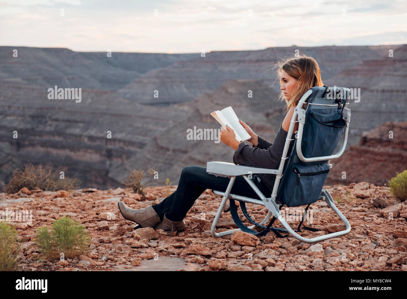Giovane donna in impostazione remota, seduti su una sedia da campeggio, lettura libro, Mexican Hat, Utah, Stati Uniti d'America Foto Stock