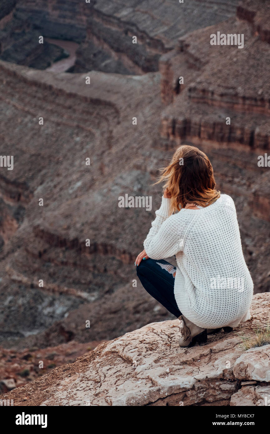 Giovane donna in impostazione remota, accovacciato sulle rocce, guardando a vista, vista posteriore, Mexican Hat, Utah, Stati Uniti d'America Foto Stock