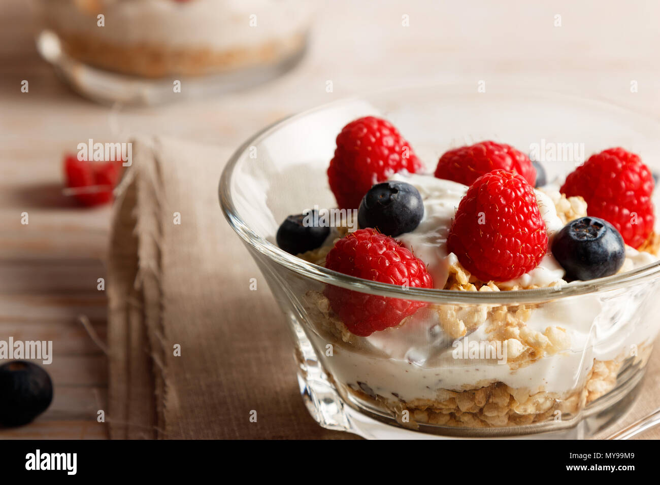 Lamponi Mirtilli, cereali e yogurt in una ciotola di vetro su un letto di sacco e di assicelle di legno. Una sana colazione per una vita sana. L'immagine orizzontale. Foto Stock