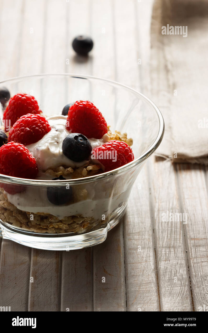 Lamponi Mirtilli, cereali e yogurt in una ciotola di vetro su assicelle di legno. Una sana colazione per una vita sana. L'immagine verticale. Foto Stock