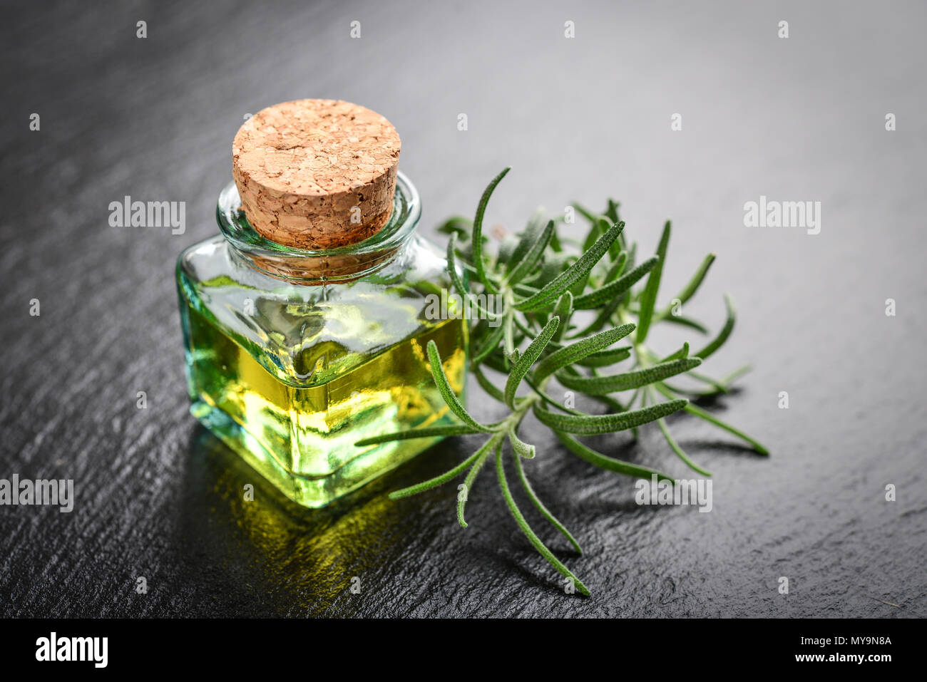 Olio essenziale di rosmarino in una bottiglia di vetro con tappo di sughero e di rosmarino fresco cantato sul nero ardesia sfondo Foto Stock