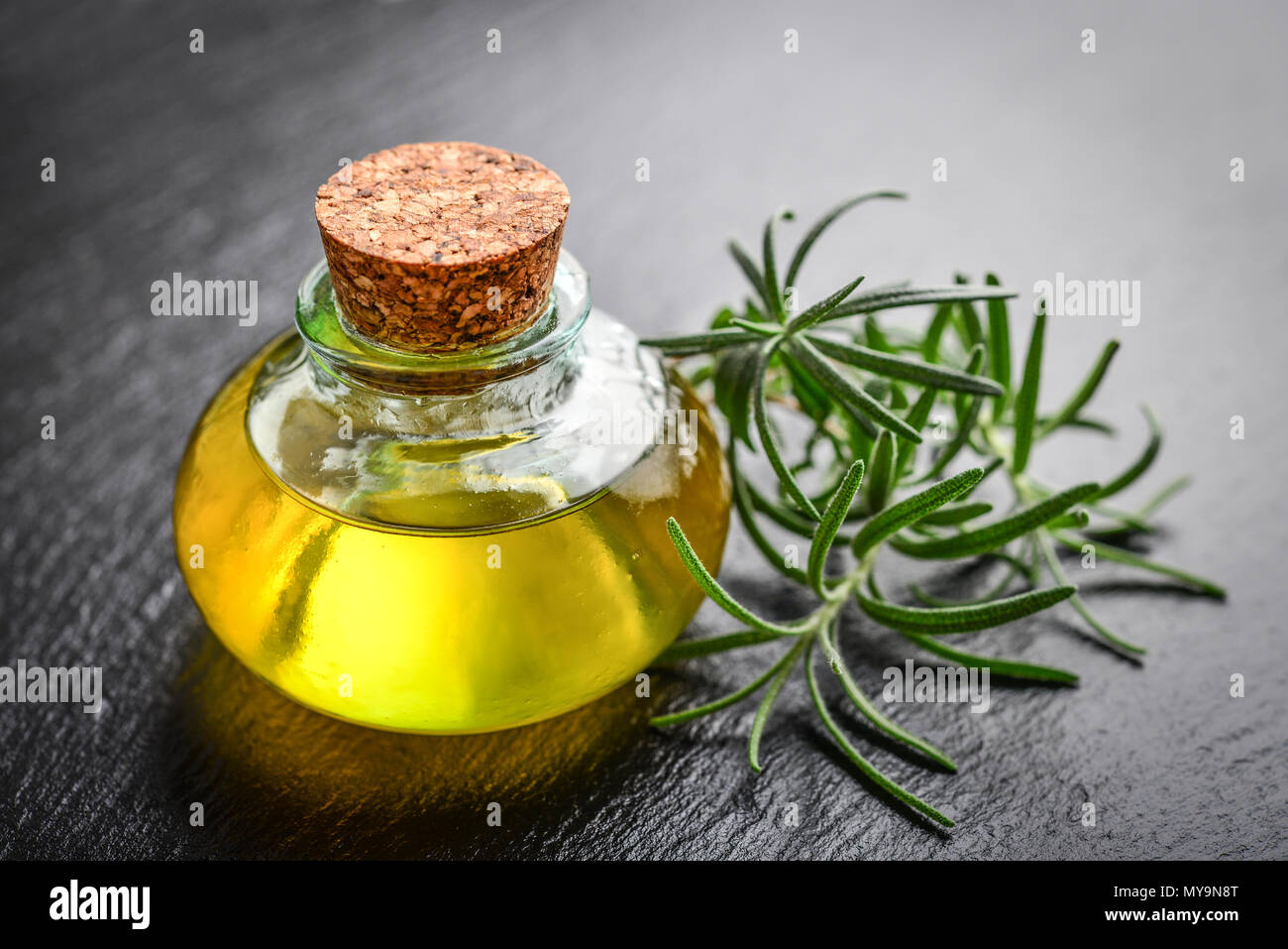 Olio essenziale di rosmarino in una bottiglia di vetro con tappo di sughero e di rosmarino fresco cantato sul nero ardesia sfondo Foto Stock