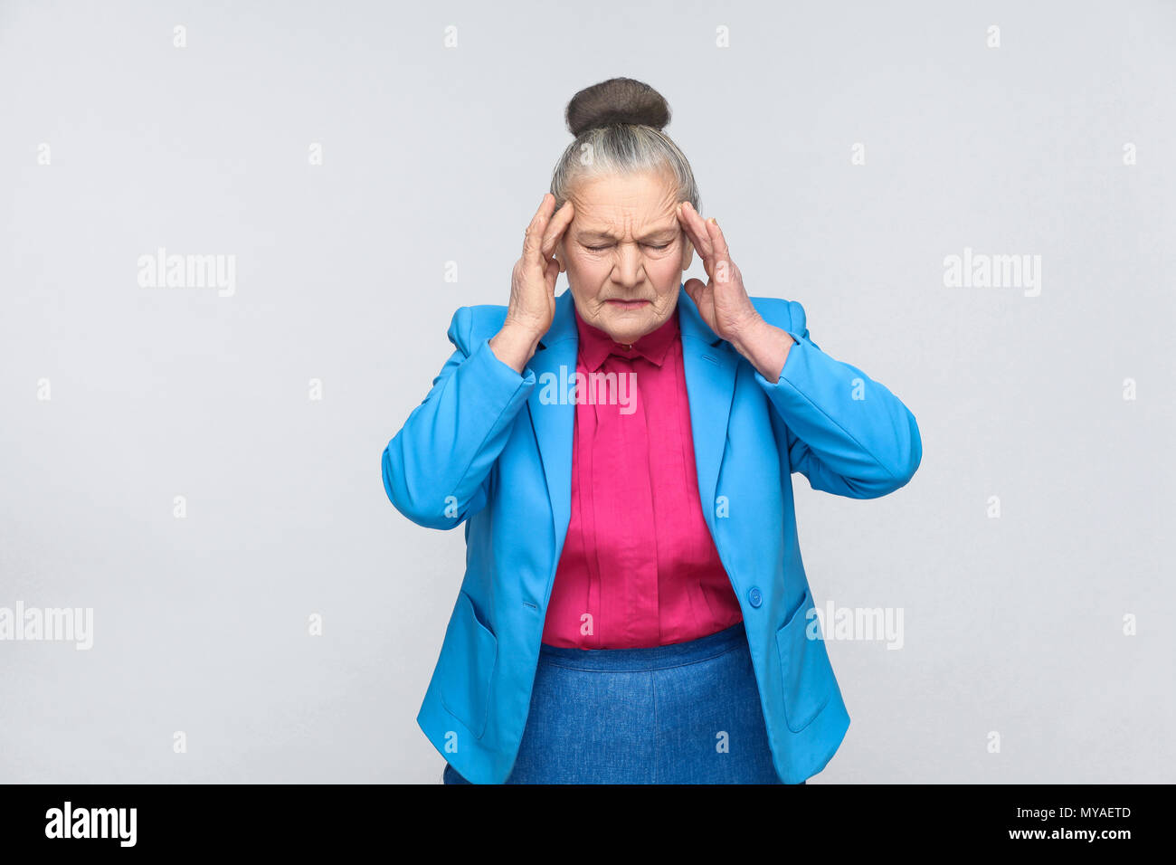 Età della donna hanno emicrania e mal di testa dolore. Portrai della nonna espressiva con la luce blu tuta e maglia rosa in piedi con raccolti bun grigio hai Foto Stock