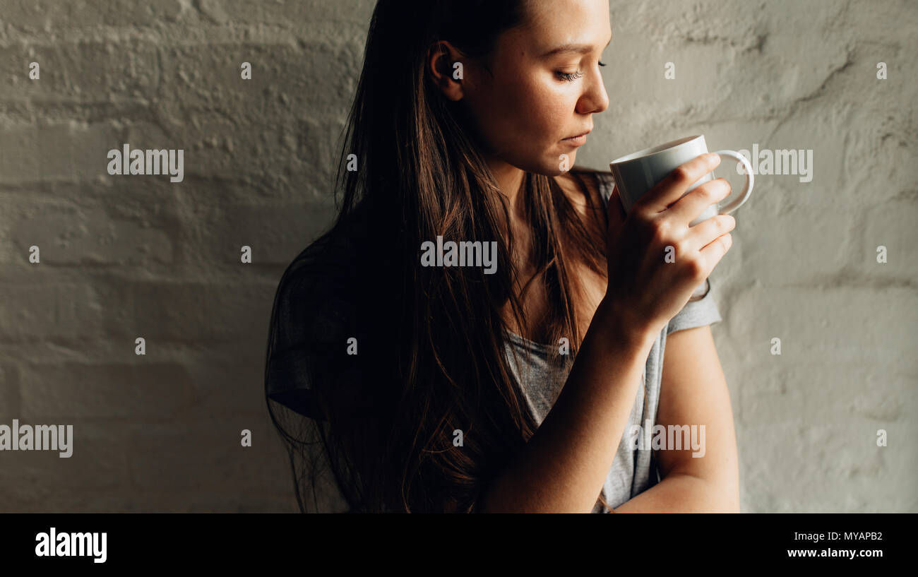 In prossimità di una donna in piedi contro la parete tenendo una tazza di caffè. Donna che guarda la tazza di caffè in mano in piedi accanto a una parete. Foto Stock