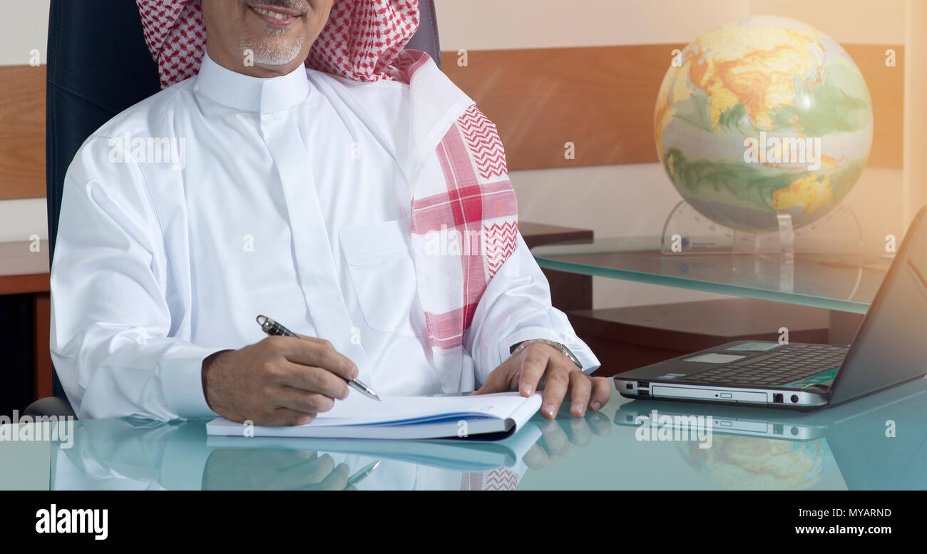 Senior imprenditore saudita la scrittura a mano alla sua scrivania con computer portatile Foto Stock