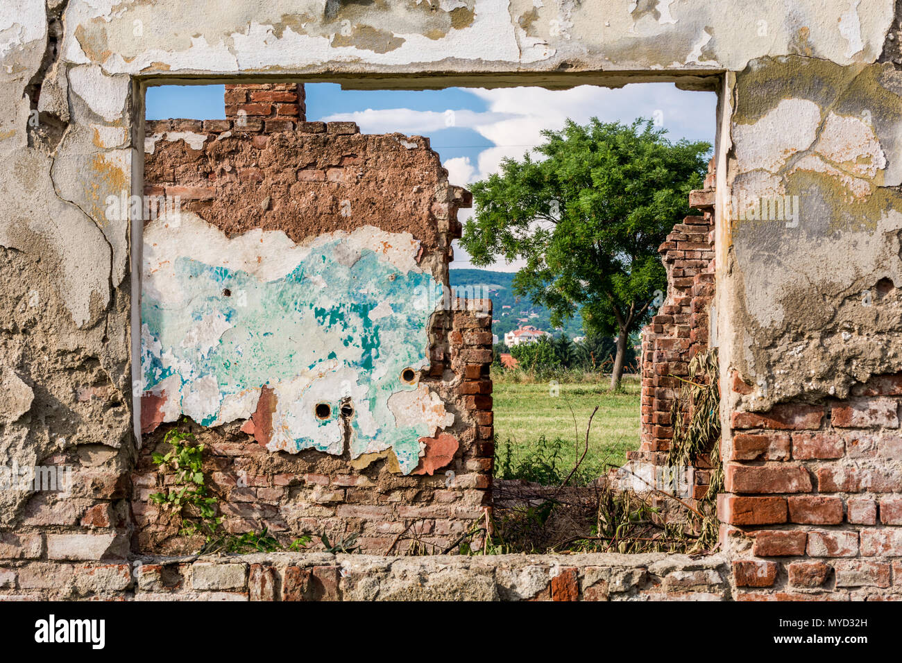 Parete danneggiata e il telaio del finestrino con vista di rovine e camera campo verde di erba, albero e il blu del cielo nuvoloso. Rosso la parete in mattoni con incrinature Foto Stock