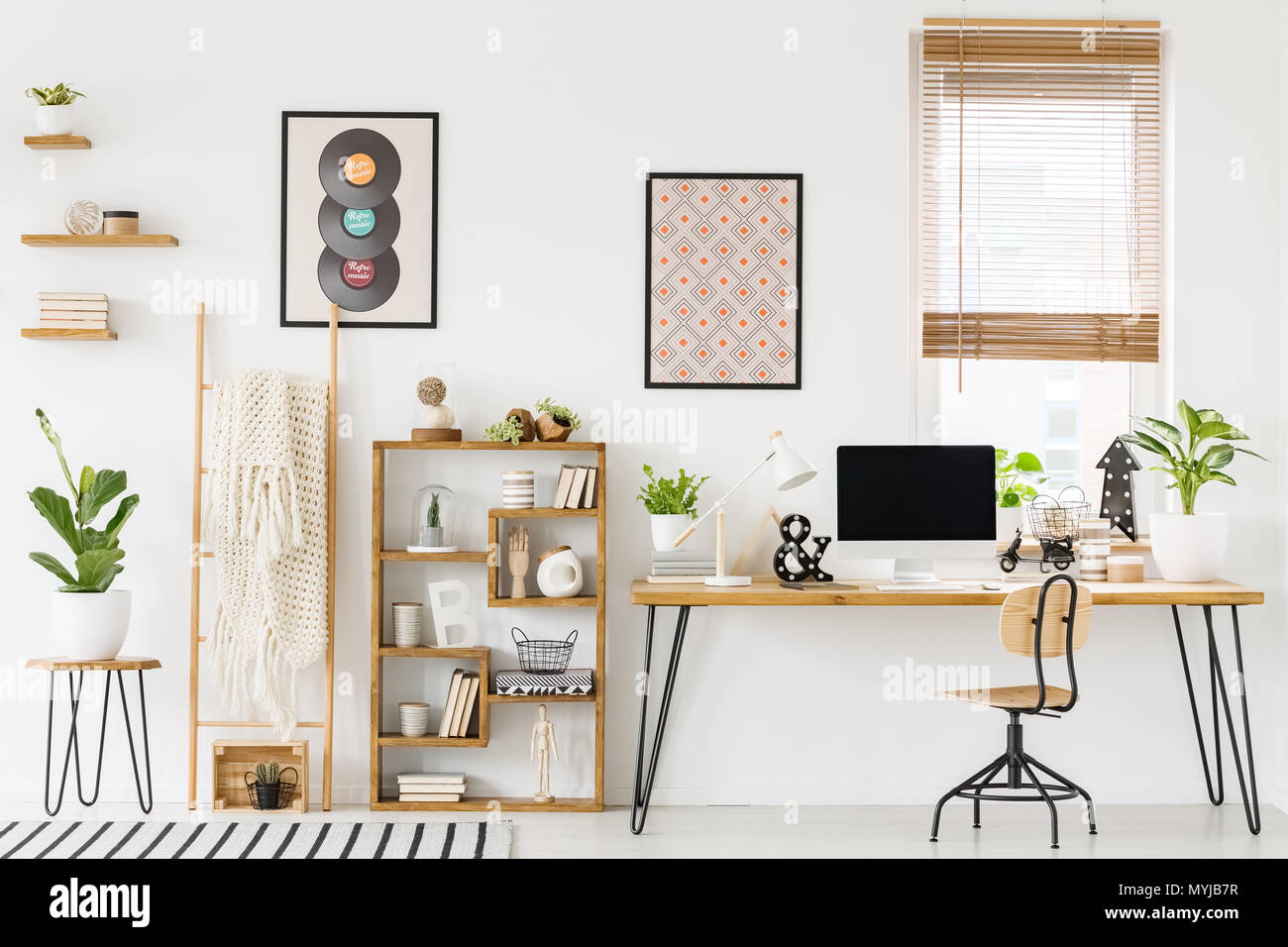 Foto reale di un luogo di lavoro interno con un computer su un tavolo accanto a una sedia e un ripiano con ornamenti, poster su una parete e una finestra con persiane Foto Stock
