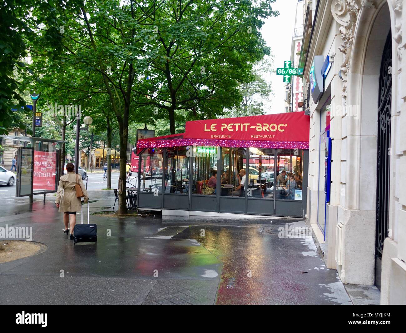 Donna che indossa un trench coat e tirando una valigia, percorrendo a piedi il marciapiede bagnato davanti a un ristorante, Montparnasse, Parigi, Francia Foto Stock