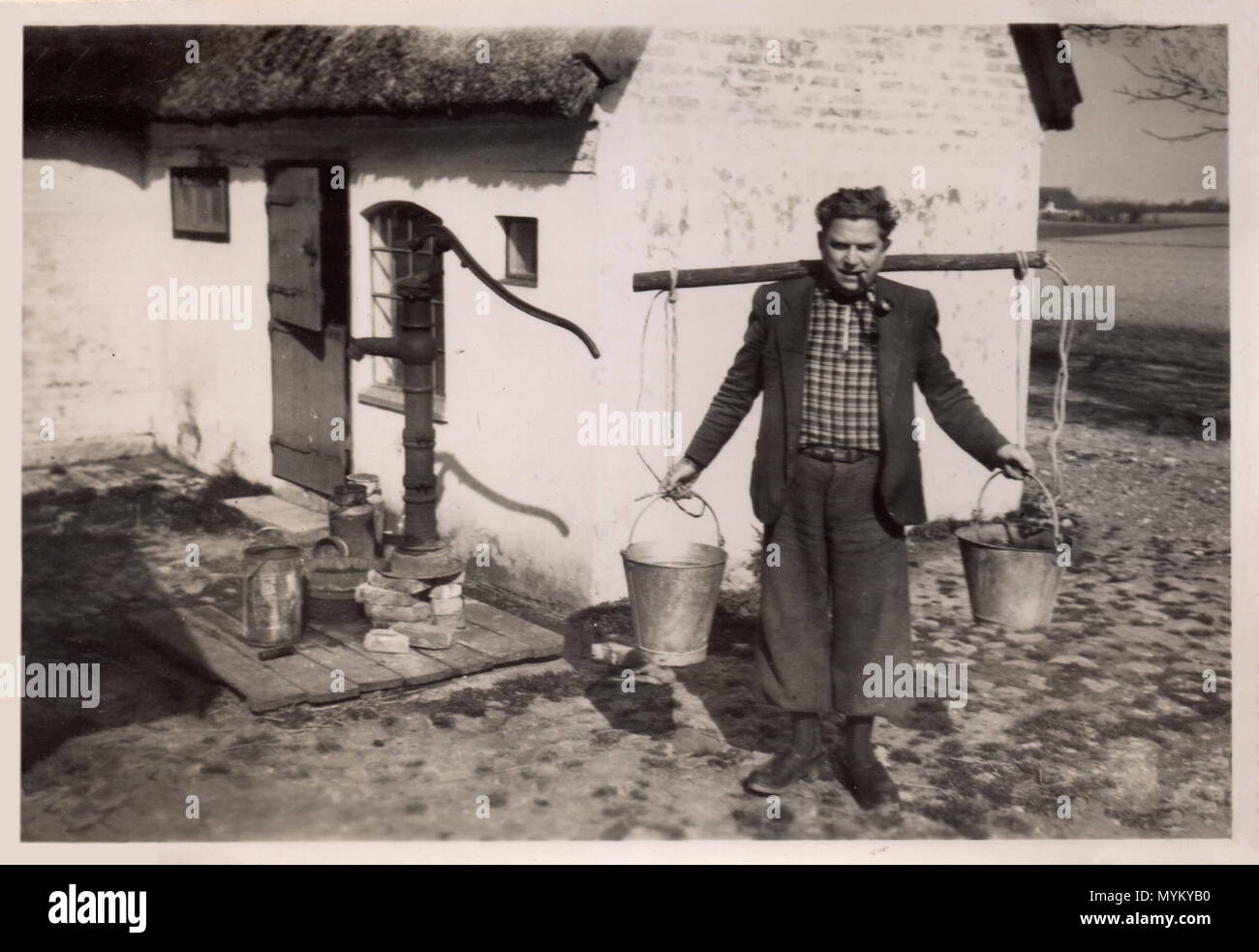 Danese un contadino, circa 1925, al di fuori del suo cottage con tetto in paglia. Egli porta due secchi da un giogo sul suo retro, probabilmente con acqua dalla vecchia pompa proprio dietro di lui; egli indossa una camicia di camicia a quadretti e un tubo, come egli sorride per il suo ritratto da adottare. Foto Stock