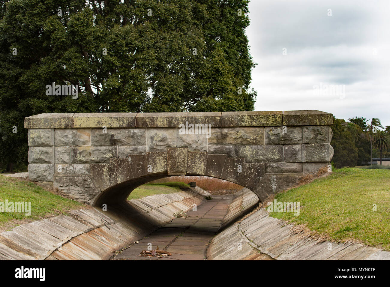 Un Romano voussoir stile arco ponte della locale pietra arenaria di Sydney abbraccia una tempesta aperto di scarico di acqua che fluisce in uno degli stagni in Centennial Park Foto Stock