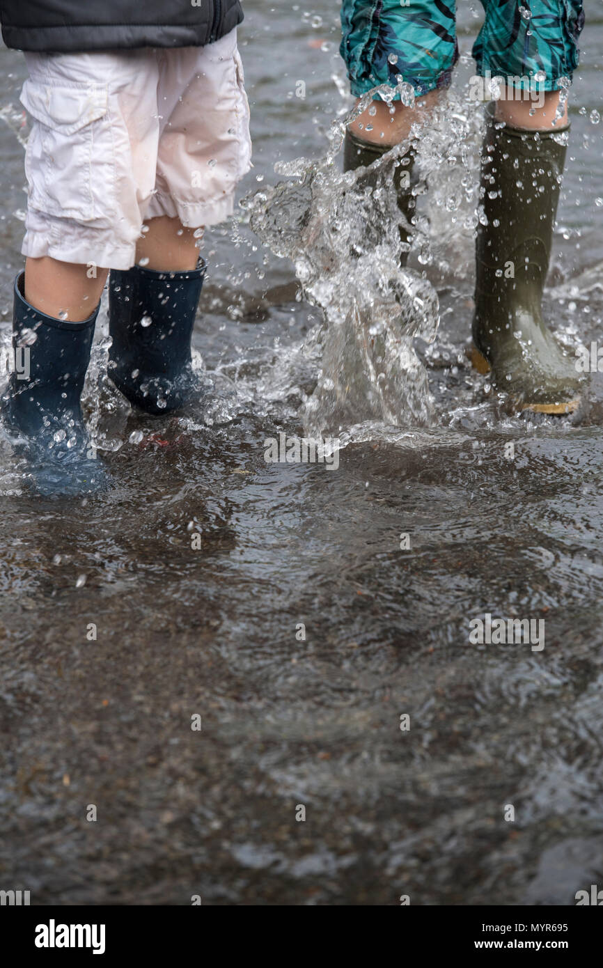Ragazzi indossando wellingtons schizzi in acqua di inondazione in una strada dopo la pioggia torrenziale Foto Stock