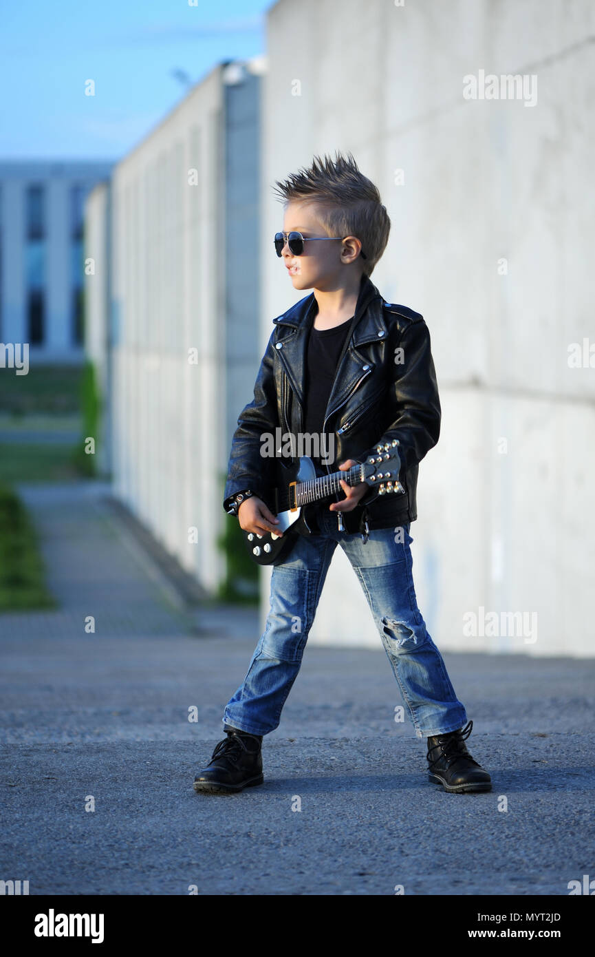 Un piccolo e grazioso ragazzo chitarrista in giacca di pelle e occhiali da  sole a suonare la chitarra. Giovani idolo. Gli interessi dei bambini e gli  hobby. Essere come una rock star