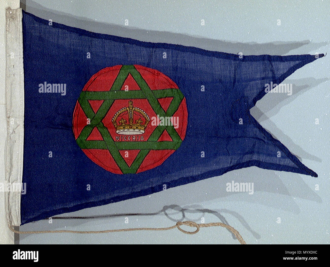 . Inglese: Ampia pennant, Direttore della Nigeria Marine ampia pennant del Direttore della Nigeria Marine. Il pavese è di lana e di fibre sintetiche e mescola il ensign ha un paranco di cotone contenente una fune di hissage il ensign. La fiamma è macchina cuciti con un disegno stampato. Design: un insegna blu con un disco rosso nel paranco, sul quale è a sei punte stella verde con una corona d'oro nel centro e NIGERIA" in lettere bianche. Indossato il capitano F. W. J. Skutil l'ultima gita ufficiale della Nigeria stazioni Marine e volato sullato sede flagstaff l'ultimo giorno prima di indipendenza. Il GRE Foto Stock