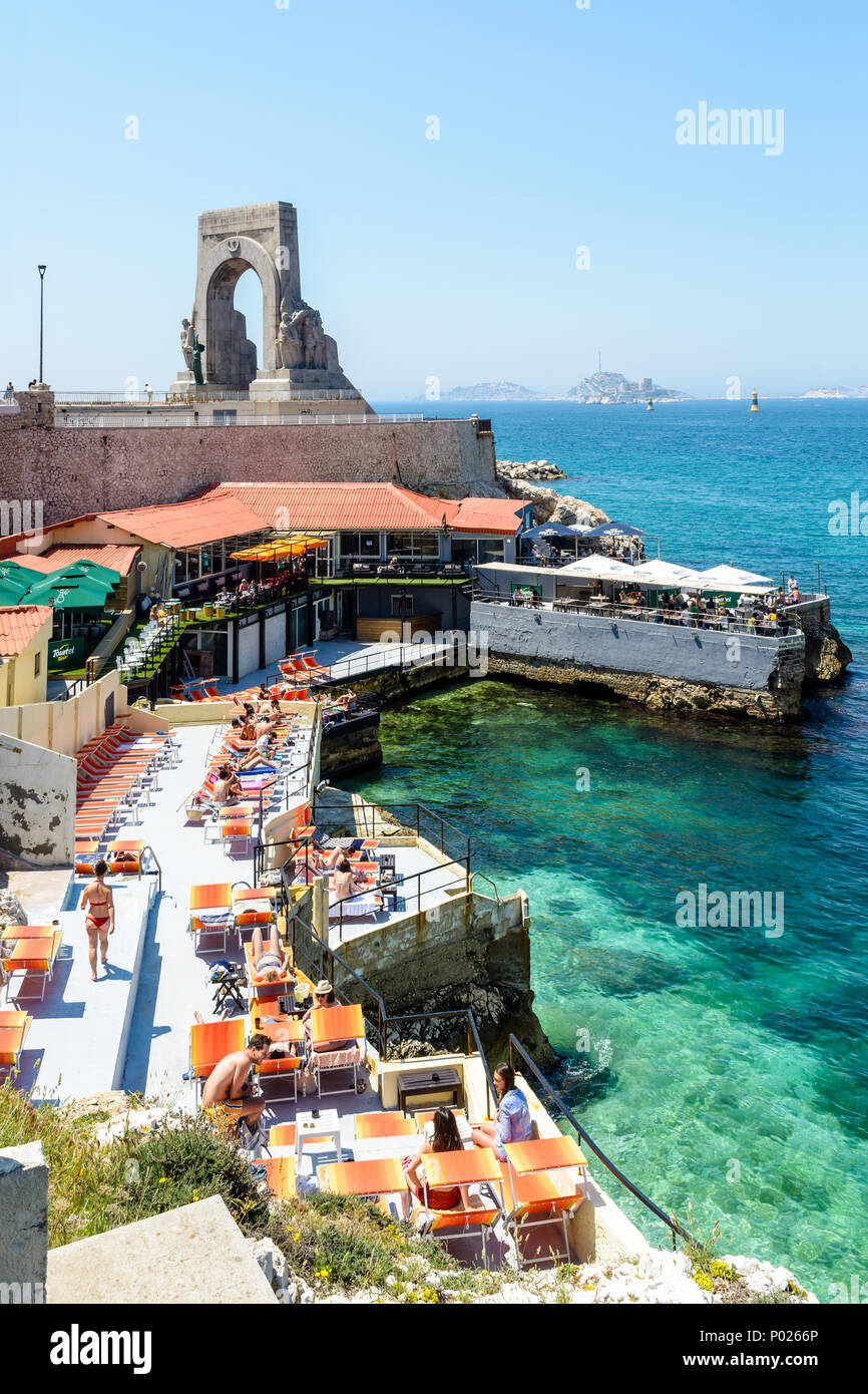 Un ristorante sulla spiaggia in riva al di sotto del memoriale di guerra per l'esercito orientale a Marsiglia, Francia, ha impostato la sua terrazza con ombrelloni e sedie a sdraio. Foto Stock