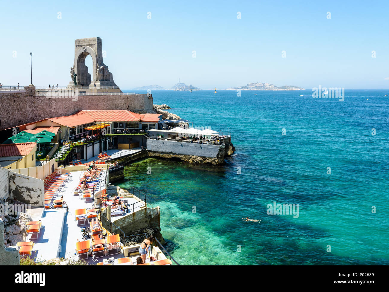 Un ristorante sulla spiaggia in riva al di sotto del memoriale di guerra per l'esercito orientale a Marsiglia, Francia, ha impostato la sua terrazza con ombrelloni e sedie a sdraio. Foto Stock