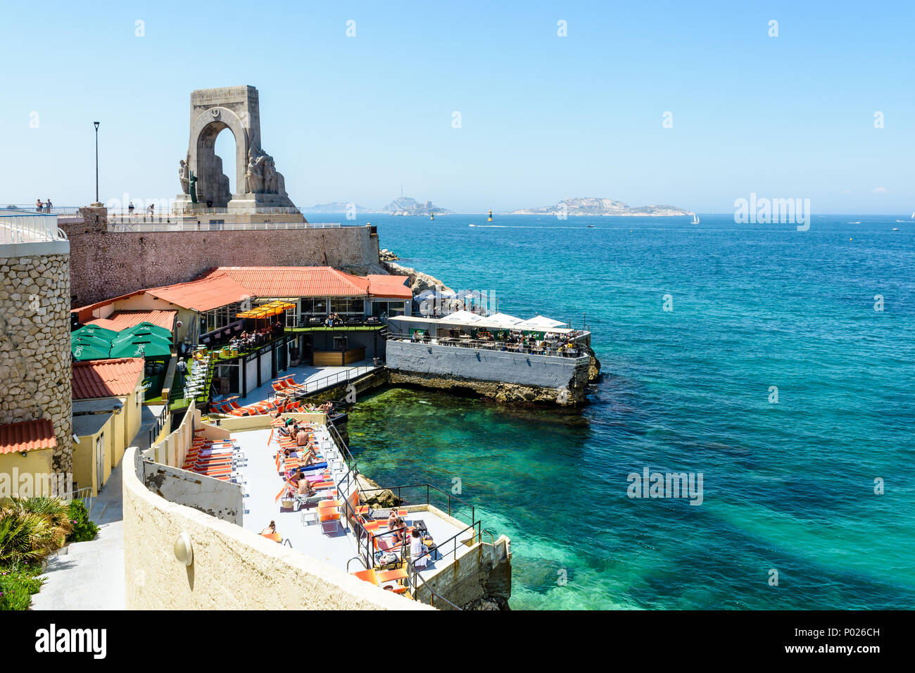 Un ristorante sulla spiaggia in riva al di sotto del memoriale di guerra per l'esercito orientale a Marsiglia, Francia, ha impostato la sua terrazza con ombrelloni e sedie a sdraio. Foto Stock