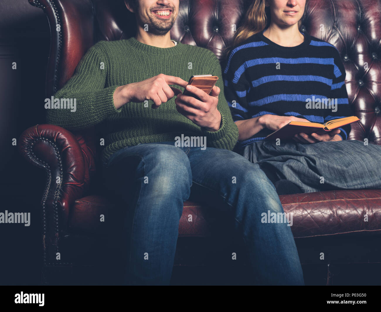 Un felice giovane uomo su un divano è utilizzando il suo smartphone accanto a una donna la lettura di un libro Foto Stock