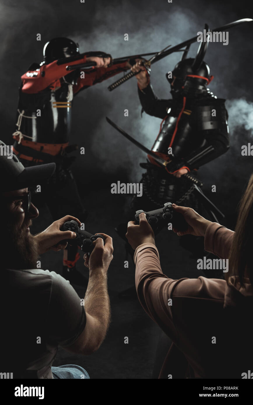 Vista posteriore di un uomo e di una donna che gioca combattimenti samurai nella vita reale con i gamepad su nero Foto Stock