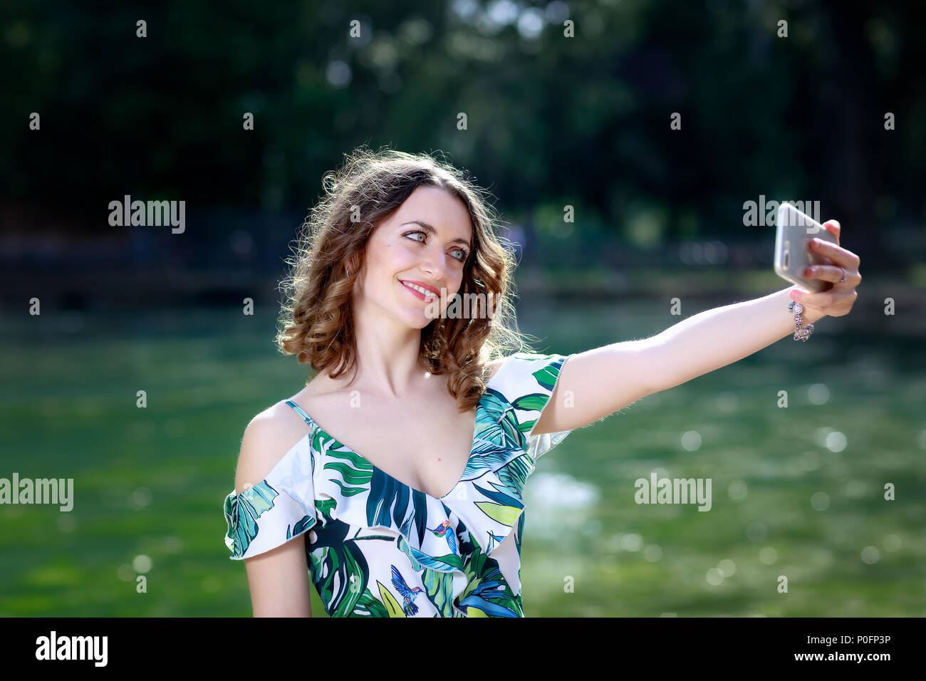 Brunette girl prendere un selfie e sorriso. Sullo sfondo il lago verde nel parco all'aperto. Una bella ragazza europea, di carnagione chiara, blue eye. Foto Stock