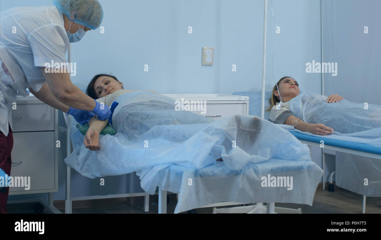 Due pazienti di sesso femminile su gocciolamenti giacente su letti di ospedale essendo controllati da due medici Foto Stock