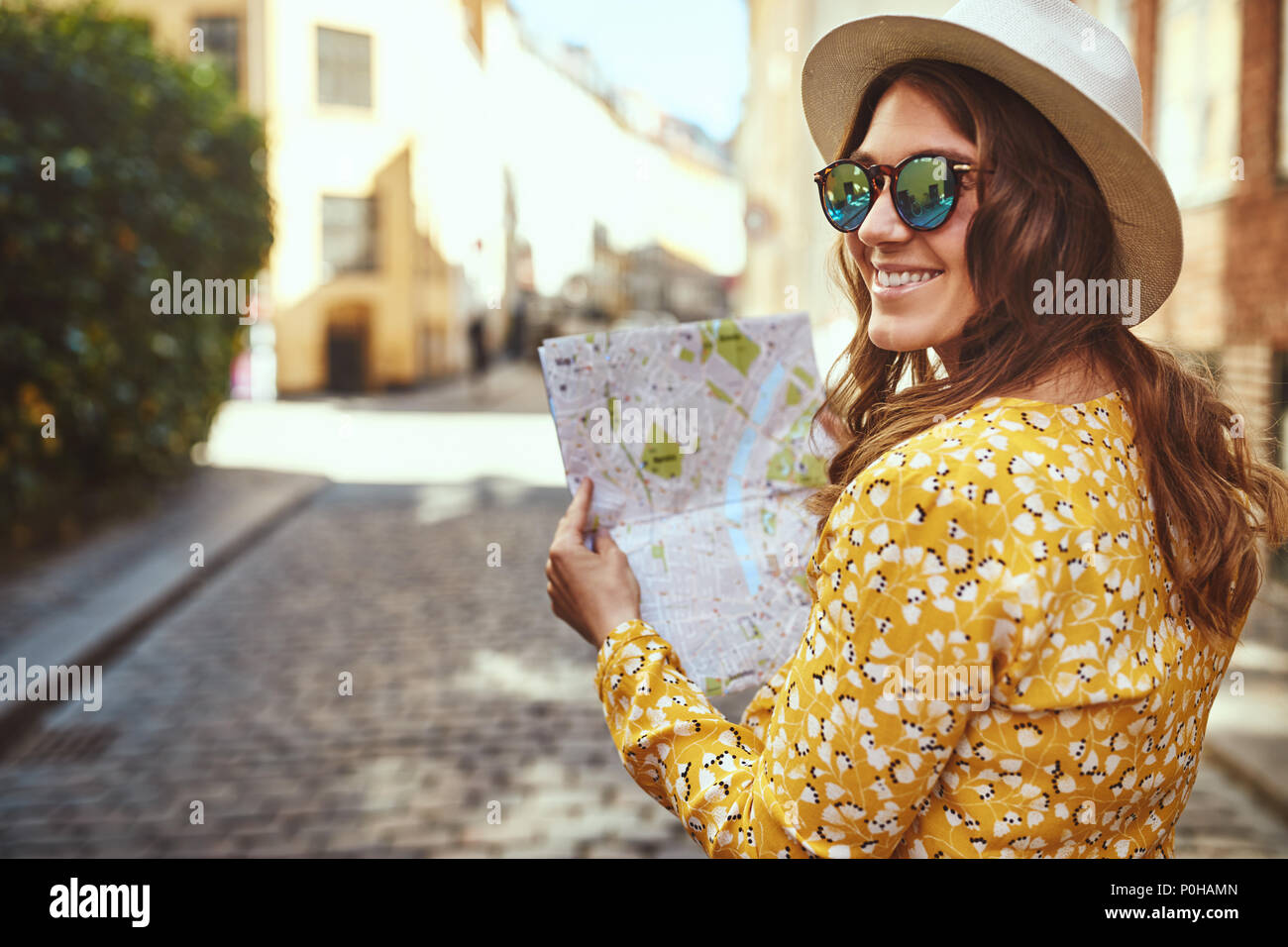 Sorridente bruna giovane donna che indossa un cappello e occhiali da sole guardando sopra la sua spalla mentre esplorare le strade di ciottoli di una città con una mappa Foto Stock