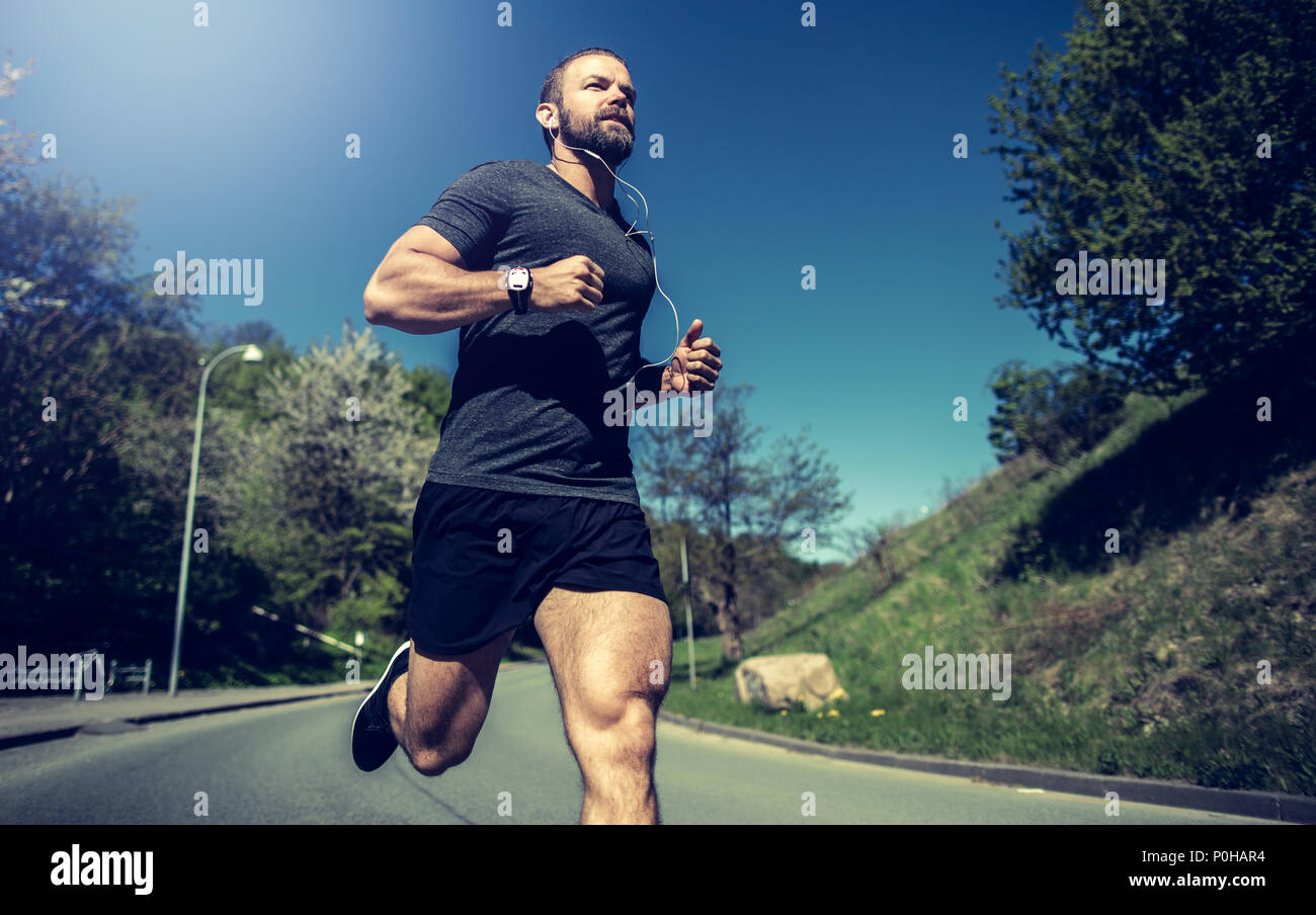 Montare il giovane uomo abbigliamento sportivo in ascolto di musica in cuffia mentre è in esecuzione da solo lungo una strada di campagna Foto Stock