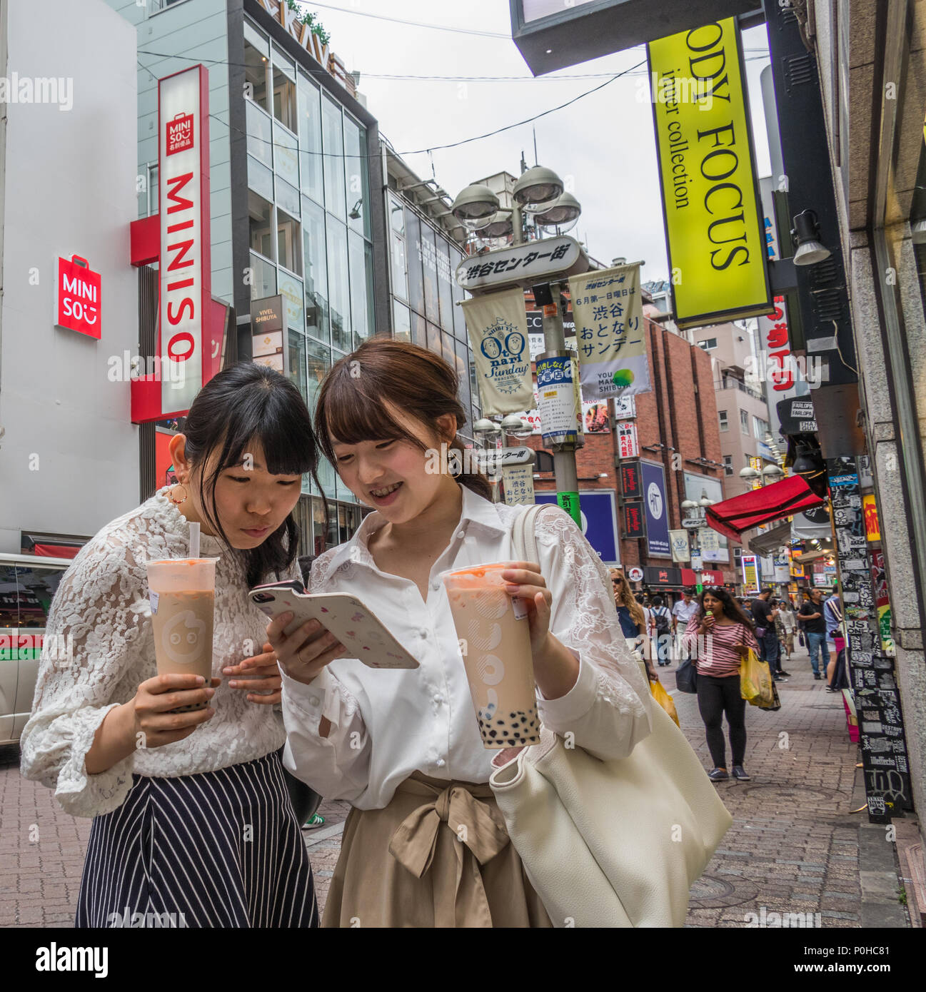 Due giovani donne, tenendo bicchieri per bevande, godetevi la lettura di un messaggio di testo sul telefono cellulare, Shibuya street, Tokyo, Giappone Foto Stock