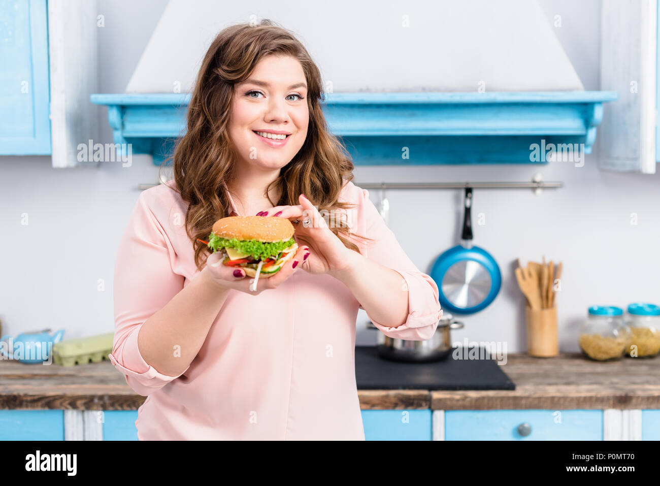 Ritratto di giovane donna sovrappeso con burger in mani in cucina a casa Foto Stock