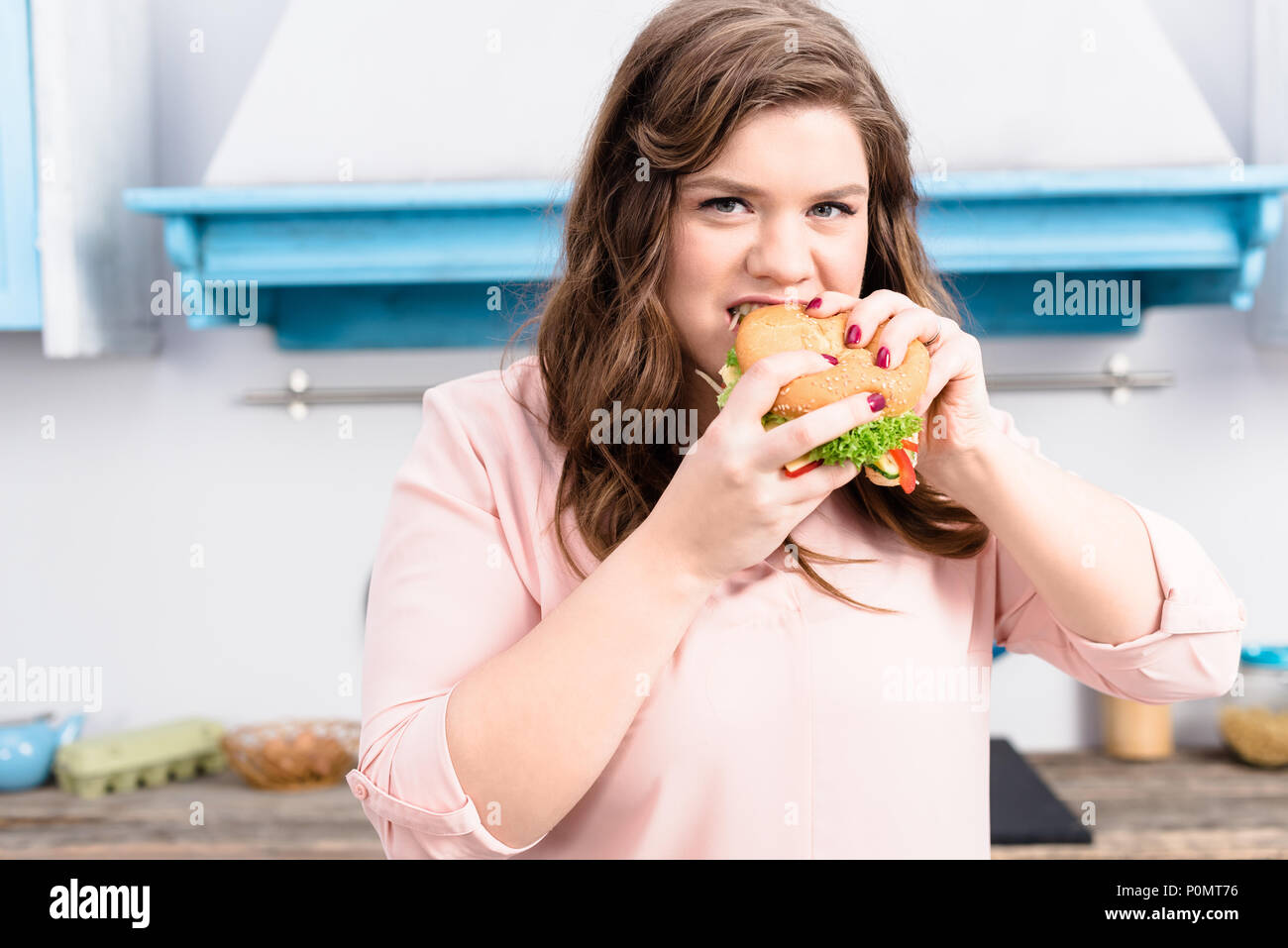 Ritratto di giovane donna sovrappeso mangiare hamburger in mani in cucina a casa Foto Stock