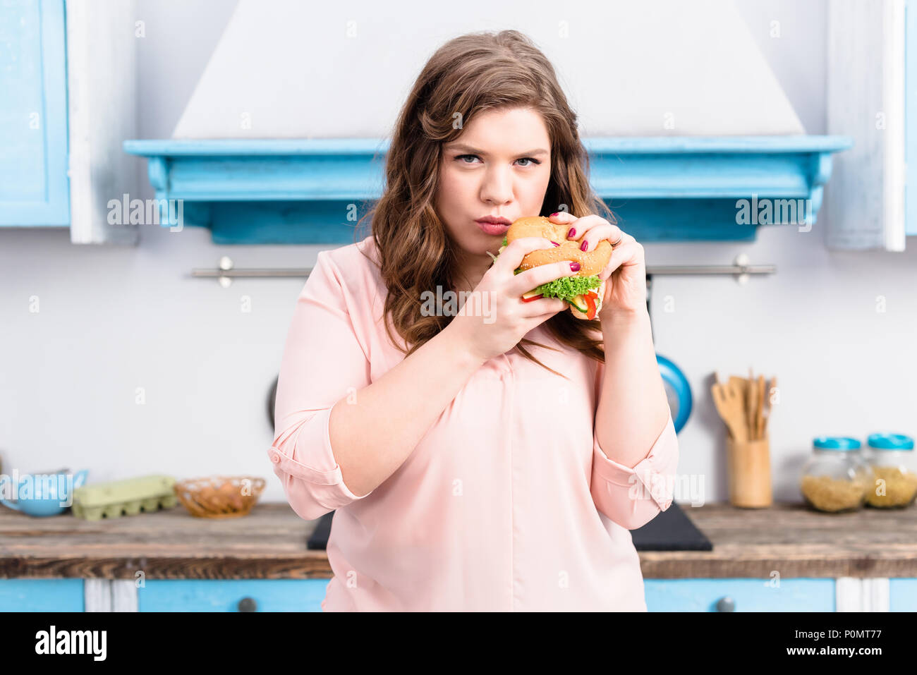 Ritratto di giovane donna sovrappeso con burger in mani in cucina a casa Foto Stock