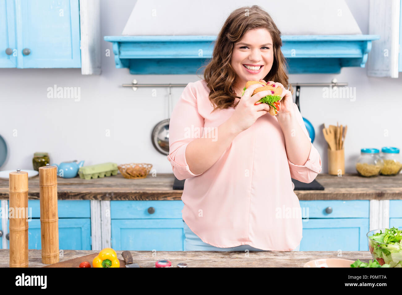 Ritratto di sovrappeso donna sorridente con il burger in mani in cucina a casa Foto Stock