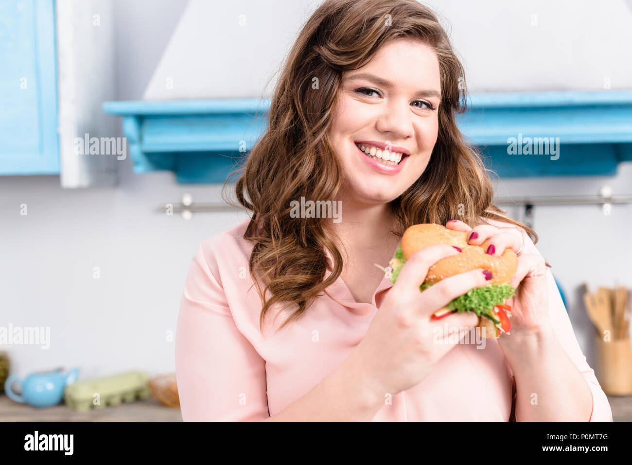 Ritratto di sovrappeso donna sorridente con il burger in mani in cucina a casa Foto Stock