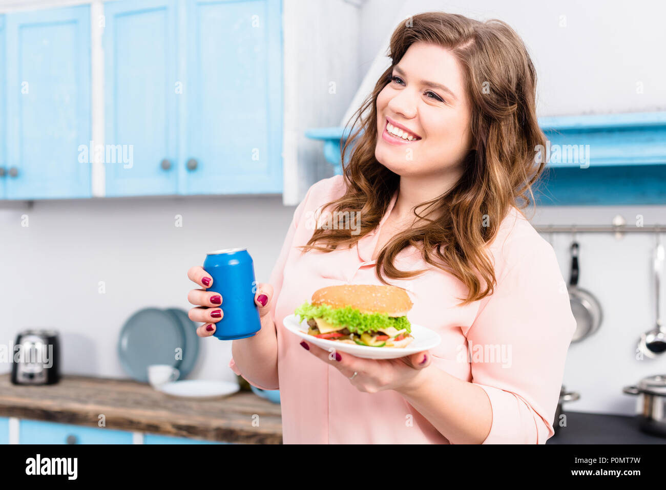 Il sovrappeso donna sorridente con soda drink e burger in mani in cucina a casa Foto Stock