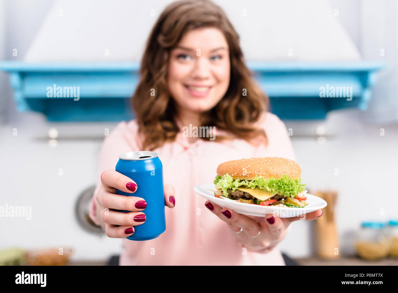 Messa a fuoco selettiva del sovrappeso donna con drink di soda e burger in mani in cucina a casa Foto Stock