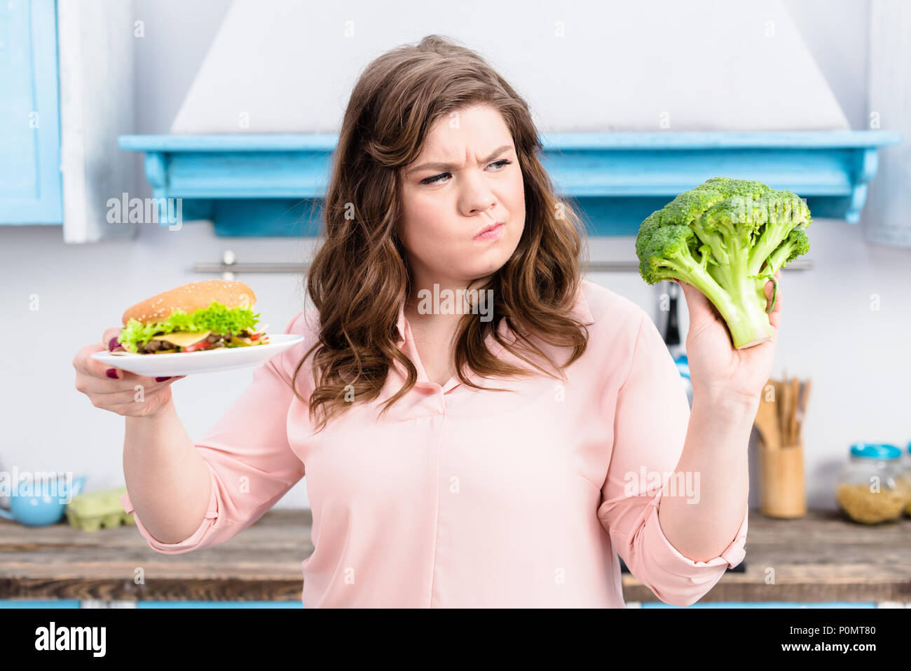 Ritratto di donna sovrappeso con burger e broccoli freschi in mani in cucina a casa, mangiare sano concetto Foto Stock