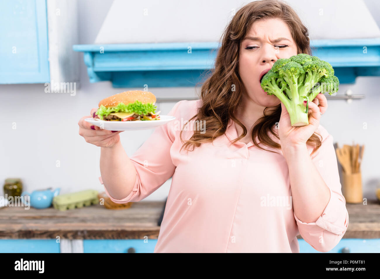 Ritratto di donna sovrappeso con burger mordere di broccoli freschi in mani in cucina a casa, mangiare sano concetto Foto Stock
