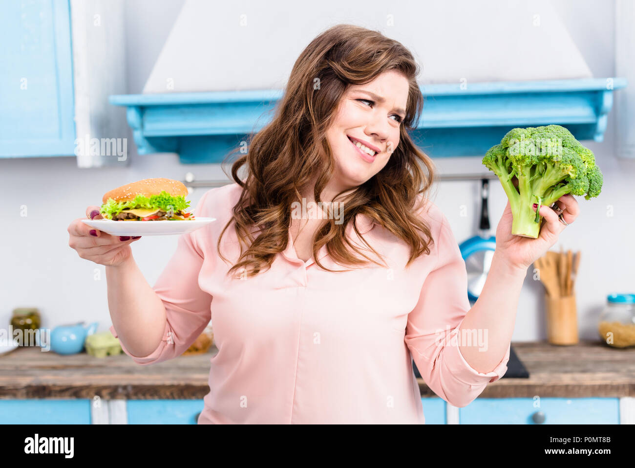 Ritratto di donna sovrappeso con burger e broccoli freschi in mani in cucina a casa, mangiare sano concetto Foto Stock