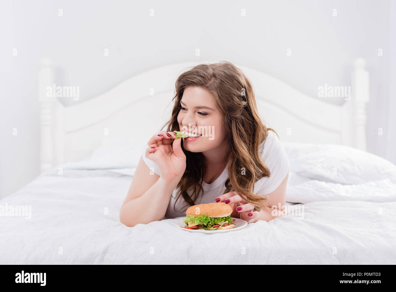 Il sovrappeso donna in pigiama mangiando burger su letto di casa Foto Stock