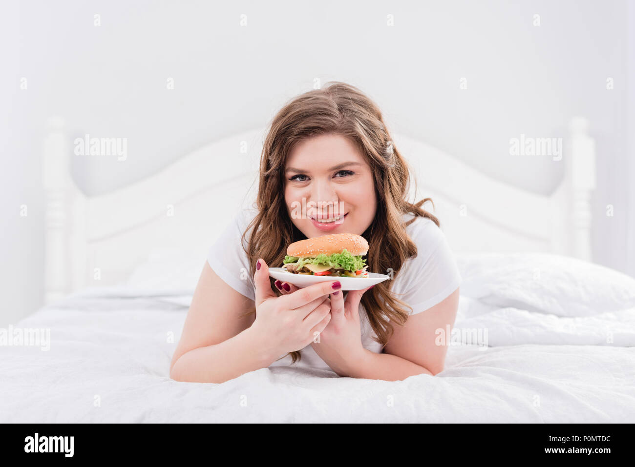 Ritratto di sovrappeso donna sorridente in pigiama con burger su letto di casa Foto Stock