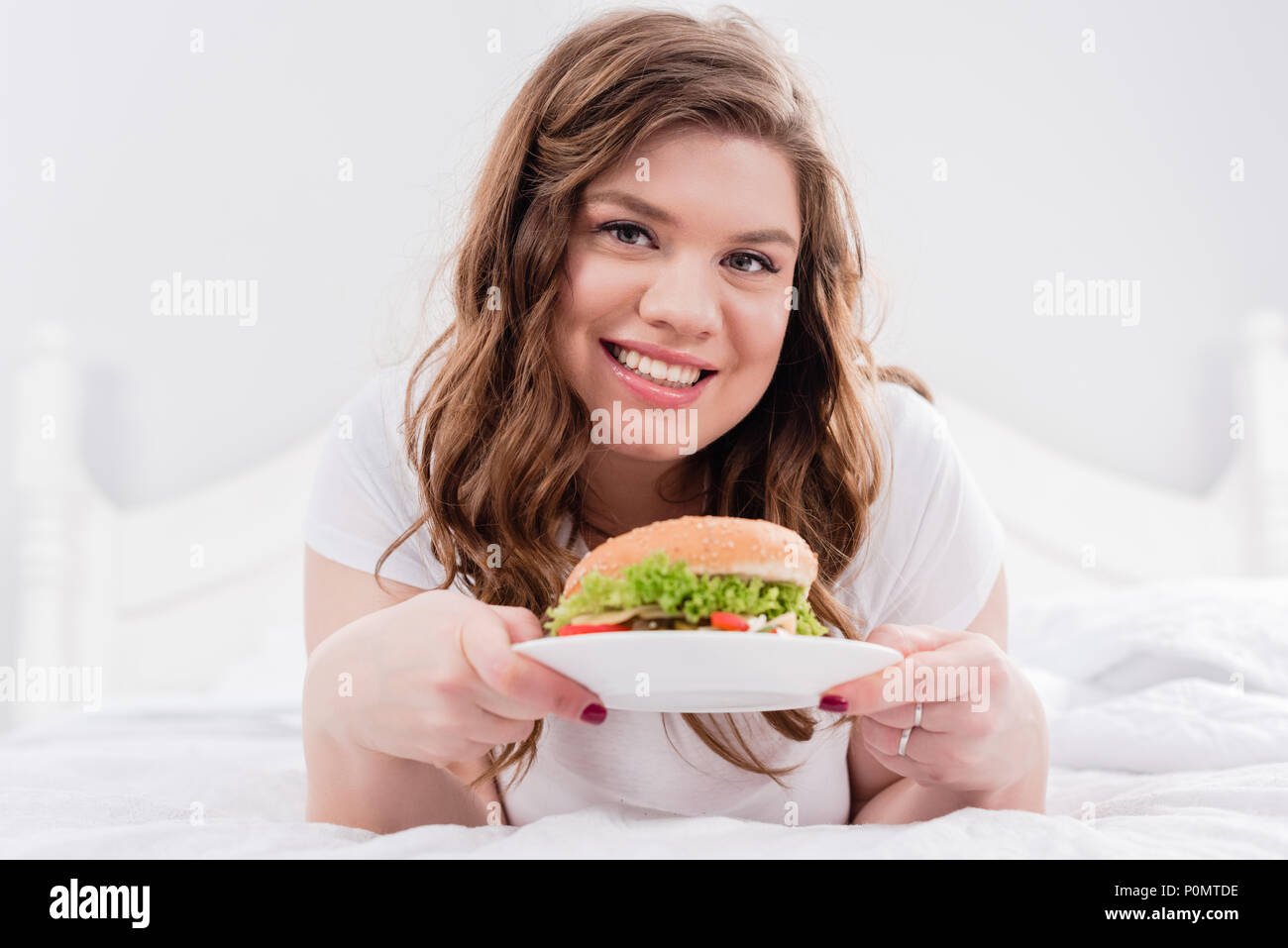 Ritratto di sovrappeso donna sorridente in pigiama con burger su letto di casa Foto Stock