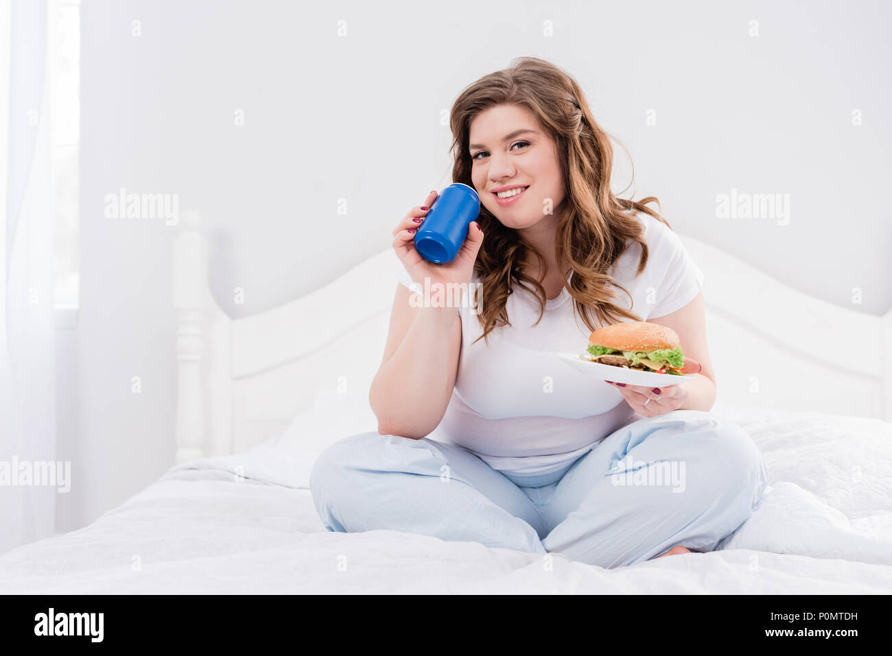Il sovrappeso donna sorridente in pigiama con soda drink e burger su letto di casa Foto Stock