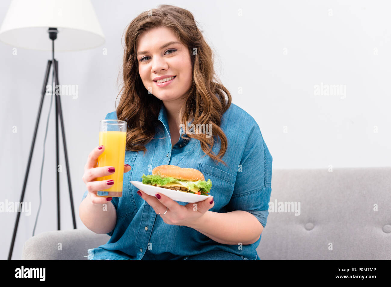 Il sovrappeso donna sorridente con un bicchiere di succo di frutta e burger sulla piastra in mani a casa Foto Stock