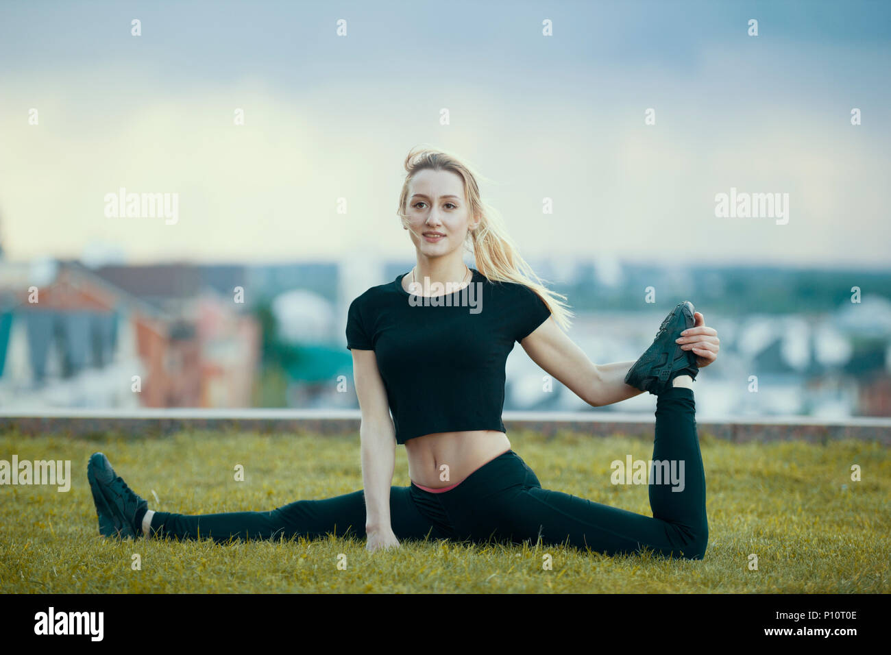 Felice giovane donna sull'erba esegue lo spago con la gamba piegata, sul paesaggio urbano di sfondo Foto Stock