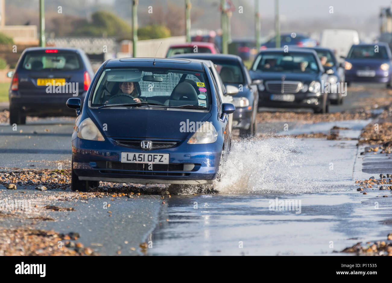 Auto guidando lentamente attraverso acqua sulla strada allagata dopo heavy rain e alta marea acqua di mare e ciottoli di spiaggia sulla strada nel West Sussex, Regno Unito. Foto Stock