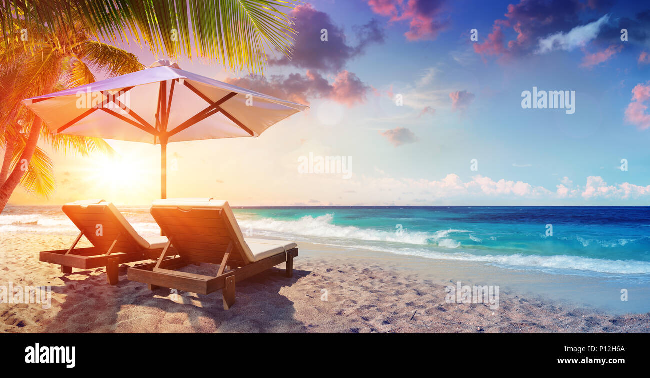 Due sedie a sdraio sotto ombrellone in spiaggia tropicale al tramonto Foto Stock